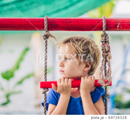 Close portrait Sad lonely freckled blue eyed cute blond little boy in t-shirt sitting on a rope ladder outside on playground in kindergarten. Daycare, activity, childhood, friendship, relationship 69726328
