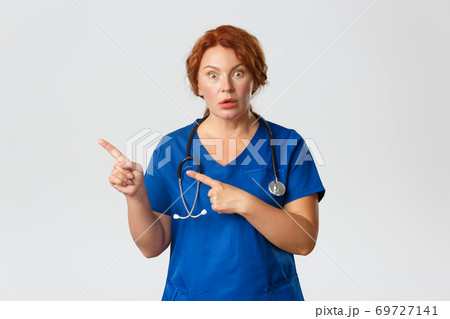 Medicine, healthcare and coronavirus concept. Portrait of shocked and impressed redhead female doctor, nurse in scrubs pointing fingers left, asking question, interested in banner, grey background 69727141