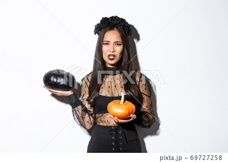 Portrait of disappointed frowning girl in witch costume, holding pumpkins and complaining, grimacing while standing over white background 69727258