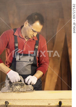 middle-aged male carpenter in googles working on workbench with electric planer 69727582