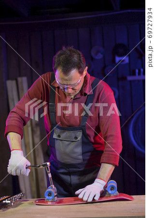 middle-aged man in jumpsuit fixing red plastic kids skateboard in home workshop 69727934