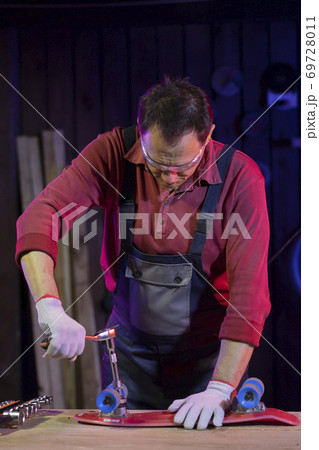 middle-aged man in jumpsuit fixing plastic kids skateboard in home workshop middle-aged man in jumpsuit fixing plastic kids skateboard in home workshop 69728011