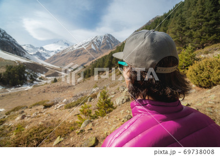 A long-haired young man in a purple down jacket and grey cap in sunglasses stands with his back to the camera and looks at a mountain gorge with snow-capped peaks in the distance 69738008