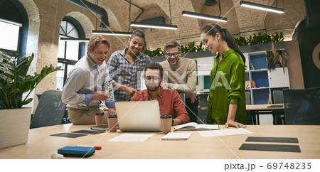 Business professionals at work. Multicultural team, group of young cheerful business people analyzing data, looking at laptop screen while working together in the office Business professionals at work. Multicultural team, group of young cheerful business people analyzing data, looking at laptop screen while working together in the office 69748235