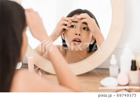 Upset asian young woman checking for pimples, looking at mirror 69749375