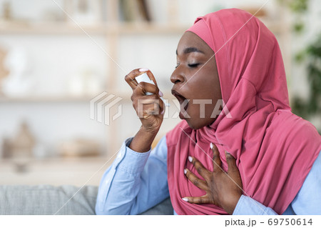 Black muslim woman in hijab using inhaler at home, suffering breathing problem 69750614