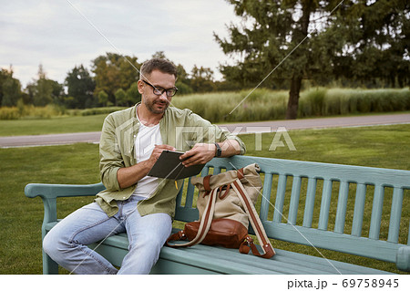 Young handsome man wearing eyeglasses sitting on the wooden bench in park and making some notes, working outdoors 69758945