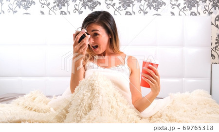 Attractive young woman gets up in bed with a cup of coffee and an alarm clock. The woman overslept 69765318