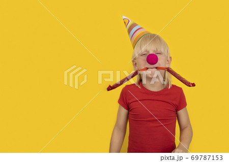 Boy blowing in festive whistle. Portrait of child with clown nose and party hat. 69787153