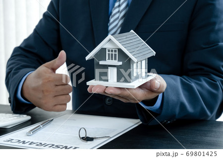 The hand of the real estate agent has a sample house with home insurance documents and keys to present to his clients who have made a purchase. Home insurance sales concept 69801425