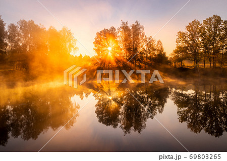 Golden misty sunrise on the pond in the autumn morning. Trees with rays of the sun cutting through it, reflected in the water. 69803265