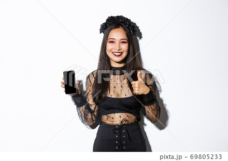 Image of happy and satisfied asian woman in halloween costume showing thumbs-up and demonstrating mobile phone screen, smiling pleased, standing over white background Image of happy and satisfied asian woman in halloween costume showing thumbs-up and demonstrating mobile phone screen, smiling pleased, standing over white background 69805233