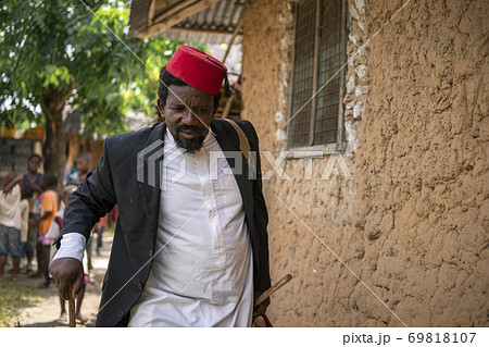 An African Older Man in Red Muslim Taqiyyah Fez Hat And Blazer on white Dress Moving a stick for lame people Near the Basic Hut with Thatched roof in Small Remote Village in Tanzania, Pemba island 69818107