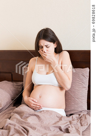 Pregnant suffering with nausea. Pregnancy symptoms, expectation toxicosis. Young vomiting woman sitting on bed. parenthood concept Pregnant suffering with nausea. Pregnancy symptoms, expectation toxicosis. Young vomiting woman sitting on bed. parenthood concept 69828618