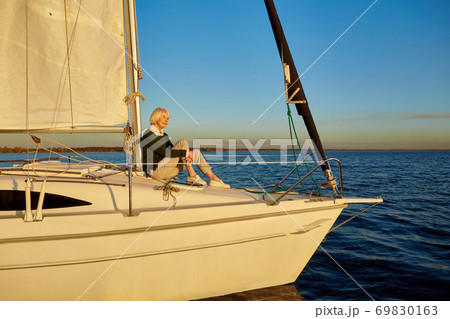 Enjoying luxury life. Beautiful senior woman sitting on the side of sailboat or yacht deck floating in the calm blue sea at sunset, looking away and smiling Enjoying luxury life. Beautiful senior woman sitting on the side of sailboat or yacht deck floating in the calm blue sea at sunset, looking away and smiling 69830163