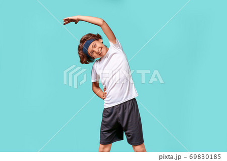 Warming up. Happy teenage boy in sportswear looking away and smiling while stretching his body before workout, standing isolated over blue background Warming up. Happy teenage boy in sportswear looking away and smiling while stretching his body before workout, standing isolated over blue background 69830185