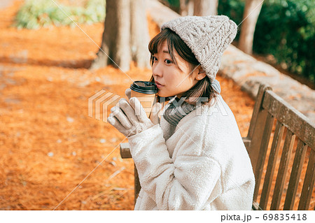 公園のベンチに座ってホットドリンクを飲む女性 69835418
