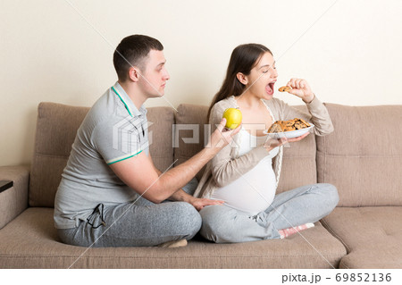 Husband tries to feed his pregnant wife with an apple but she wants cookies sitting on the sofa at home. Unhealthy diet during pregnancy concept 69852136