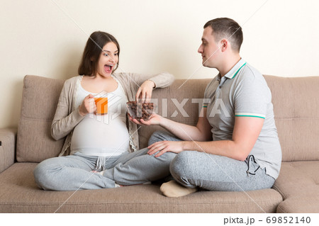 Happy husband is feeding his pregnant wife with chocolate cereal balls and juice relaxing on the sofa at home. Future father takes care about expecting mother Happy husband is feeding his pregnant wife with chocolate cereal balls and juice relaxing on the sofa at home. Future father takes care about expecting mother 69852140