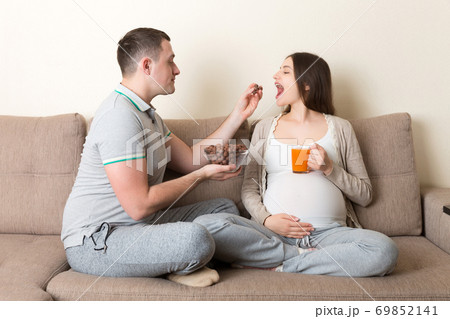Happy husband is feeding his pregnant wife with chocolate cereal balls and juice relaxing on the sofa at home. Future father takes care about expecting mother Happy husband is feeding his pregnant wife with chocolate cereal balls and juice relaxing on the sofa at home. Future father takes care about expecting mother 69852141