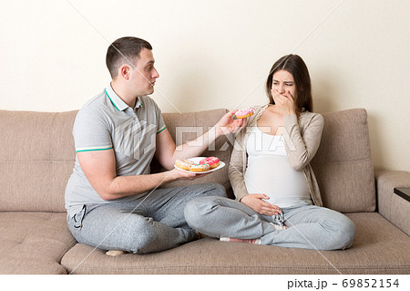 Husband offers donuts to his pregnant wife but she refuses and makes stop gesture because she feels sick. Feeling bad during pregnancy concept Husband offers donuts to his pregnant wife but she refuses and makes stop gesture because she feels sick. Feeling bad during pregnancy concept 69852154