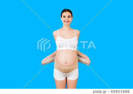 Portrait of pregnant woman in underwear putting on supporting bandage for reducing backache at blue background with copy space. Orthopedic abdominal support belt concept Portrait of pregnant woman in underwear putting on supporting bandage for reducing backache at blue background with copy space. Orthopedic abdominal support belt concept 69855886