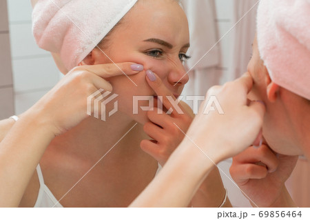 The girl squeezes out a pimple, looking in the mirror, in the bathroom. 69856464
