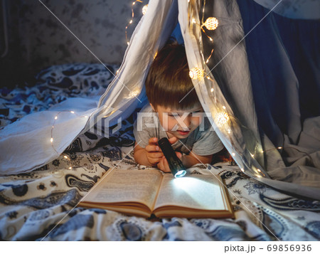 Little boy reads book with pocket light. Toddler plays in tent made of linen sheet on bed. Cozy evening with favorite book. 69856936