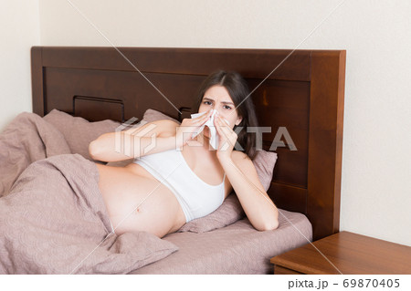 Portrait of young unhappy pregnant woman leaning on pillows on bed and blowing her nose into tissue. Future mom caught cold and sneezing Portrait of young unhappy pregnant woman leaning on pillows on bed and blowing her nose into tissue. Future mom caught cold and sneezing 69870405