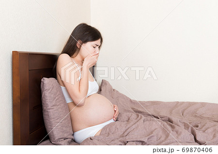 Pregnant suffering with nausea. Pregnancy symptoms, expectation toxicosis. Young vomiting woman sitting on bed. parenthood concept Pregnant suffering with nausea. Pregnancy symptoms, expectation toxicosis. Young vomiting woman sitting on bed. parenthood concept 69870406