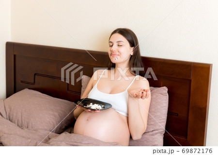 Hungry excited pregnant woman resting in bed is eating greedily a piece of cake and has a dirty mouth. Expecting mother can't stop eating 69872176