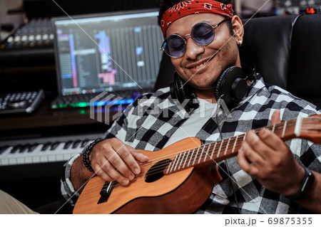 Close up portrait of happy young man, male artist in sunglasses smiling while playing ukulele, sitting in recording studio 69875355