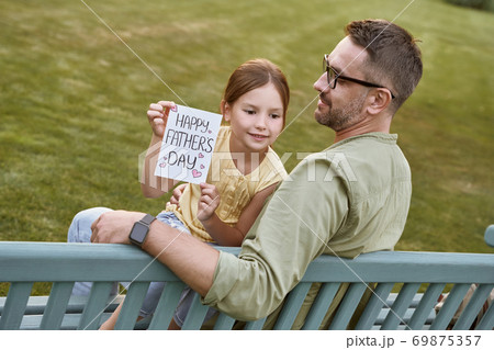 Celebrating Happy Fathers Day outdoors. Young loving father sitting on the wooden bench in park with his cute little daughter, happy girl holding handmade postcard for daddy 69875357