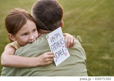 Cute little smiling girl, small daughter hugging her father and giving him handmade postcard, greeting with Happy Fathers Day while spending time together in park on a warm day Cute little smiling girl, small daughter hugging her father and giving him handmade postcard, greeting with Happy Fathers Day while spending time together in park on a warm day 69875360