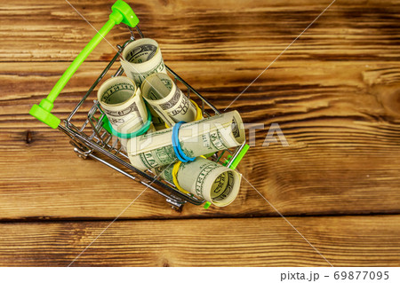 Small shopping cart with rolls of one hundred dollar bills on wooden background. Consuming concept 69877095