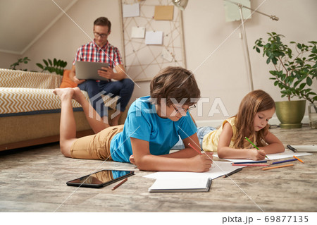 Cute little boy and girl lying on the wooden floor and drawing with colorful pencils while their father sitting on sofa and working on laptop Cute little boy and girl lying on the wooden floor and drawing with colorful pencils while their father sitting on sofa and working on laptop 69877135
