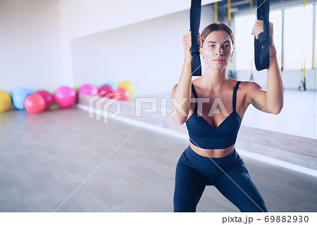 Beautiful young woman training with suspension trainer sling or suspension straps in gym. Upper body exercise concept on TRX Beautiful young woman training with suspension trainer sling or suspension straps in gym. Upper body exercise concept on TRX 69882930