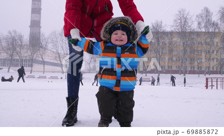 Mother leads the hands of a small smiling baby in the snow in the park. The first steps of the child Mother leads the hands of a small smiling baby in the snow in the park. The first steps of the child 69885872