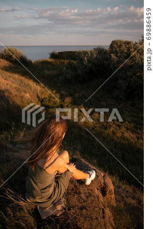 View from the back of a girl in a shirt sitting on a hill and admiring the beautiful view and sunrise or sunset 69886459