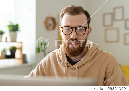 Young bearded freelance designer in eyeglasses looking at laptop display Young bearded freelance designer in eyeglasses looking at laptop display 69891300