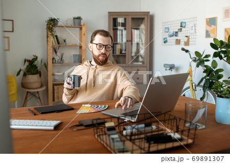 Young bearded freelancer looking at laptop display while having coffee by table Young bearded freelancer looking at laptop display while having coffee by table 69891307