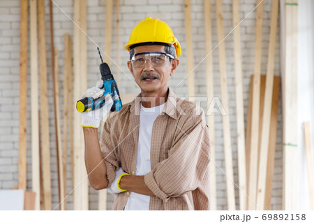 Asian senior man with hardhat and safety goggles hold electric drill in carpentry workshop. Concept senior people doing hobby after retirement. 69892158