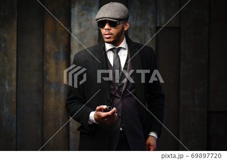 Studio shot of stylish african american man wearing coat, cap, glasses and holding smoking pipe 69897720