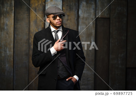 Studio shot of stylish african american man wearing coat, cap, glasses and holding smoking pipe 69897725