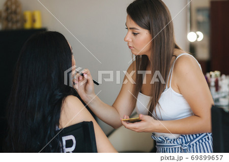 Makeup artist work in her beauty studio. Portrait of Woman applying makeup. Professional make up master. Beautiful make up artist start making a makeup for brunette woman Makeup artist work in her beauty studio. Portrait of Woman applying makeup. Professional make up master. Beautiful make up artist start making a makeup for brunette woman 69899657