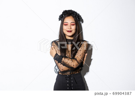 Happy cute asian woman hugging herself and smiling dreamy, wearing halloween costume of witch or widow, standing over white background 69904387