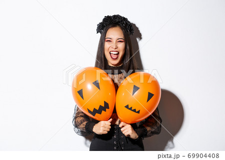 Image of asian girl in evil witch costume holding two orange balloons with scary faces, celebrating halloween, standing over white background Image of asian girl in evil witch costume holding two orange balloons with scary faces, celebrating halloween, standing over white background 69904408