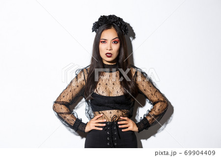Angry and pissed-off asian female magician, evil witch in black dress and wreath looking mad at someone, frowning and looking judgemental, standing over white background 69904409