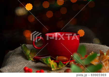 red cup with tea on saucer on bokeh background with oak acorns, autumn leaves and rowan berries 69905969