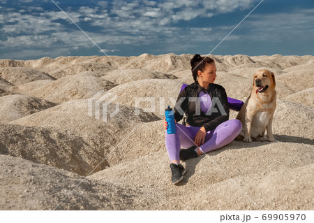 woman in sportswear sits on yellow dunes with dog labrador retriever looks each other on sunny day, copy space, concept World pet day woman in sportswear sits on yellow dunes with dog labrador retriever looks each other on sunny day, copy space, concept World pet day 69905970